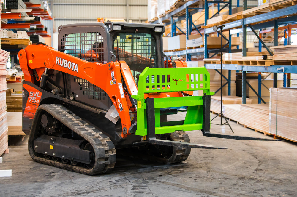 An orange Kubota SVL75-2 skid steer loader equipped with a green Impact pallet fork attachment inside a warehouse. The loader is positioned between rows of shelving filled with wooden materials, ready for material handling tasks.