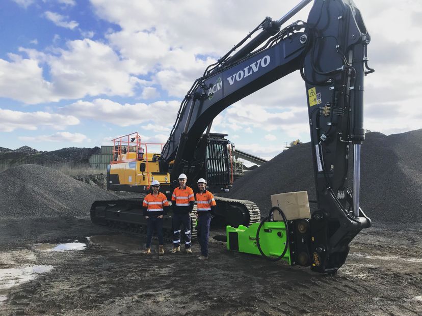 Three construction workers in high-visibility orange uniforms and white helmets standing in front of a large black Volvo excavator with a bright green attachment. The excavator is set against a backdrop of gravel piles and a partly cloudy sky