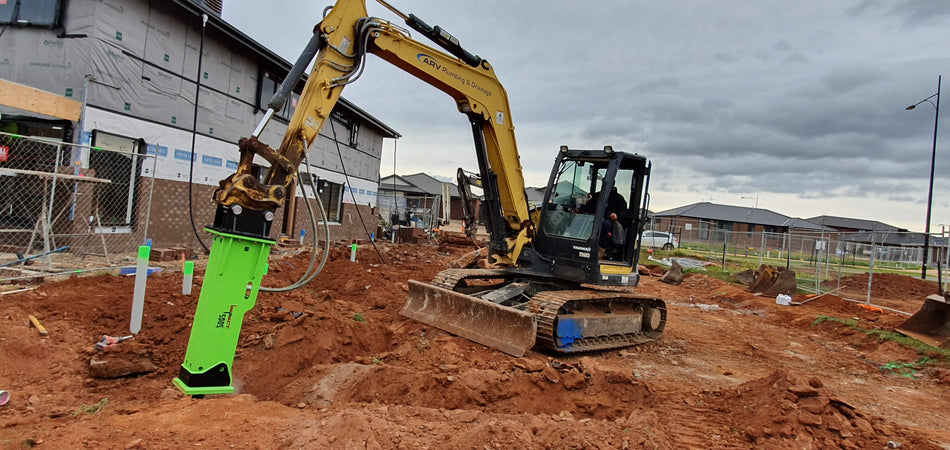 Yanmar excavator equipped with an Impact hydraulic rock breaker on a residential construction site, breaking through compacted earth. The machinery is operated by a worker from ARV Plumbing & Drainage, showcasing the durability and effectiveness of the Impact hydraulic breaker in construction and excavation tasks.