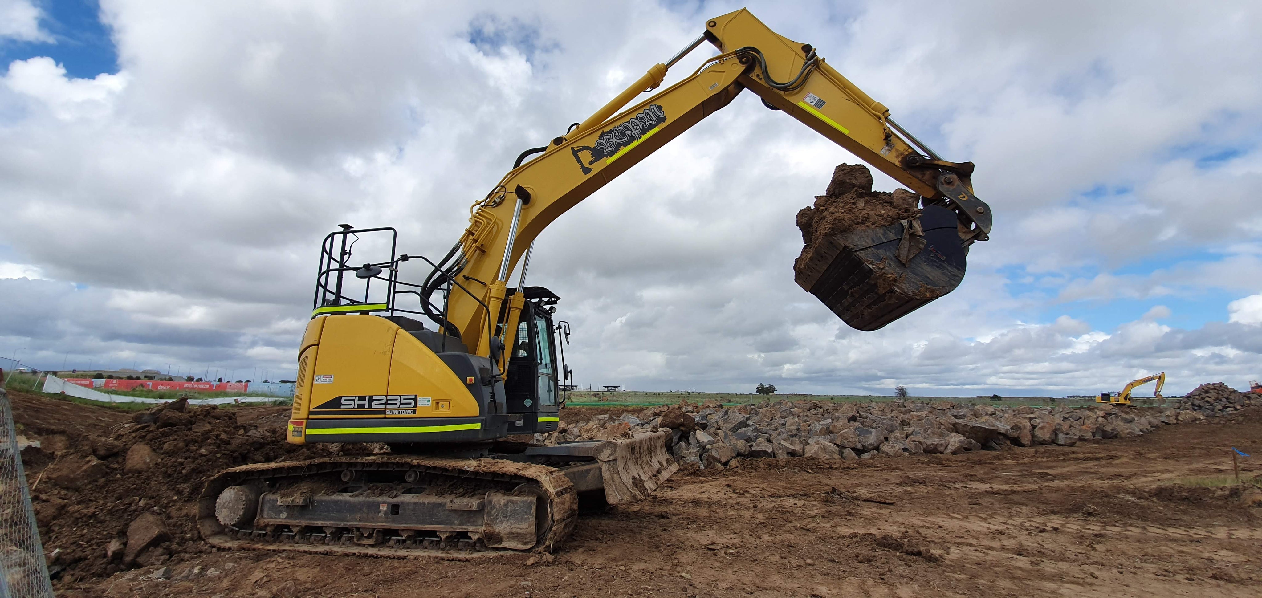 Excavator equipped with an Impact hydraulic bucket attachment lifting a load of earth on a construction site. The robust bucket, attached to a SH235 machine, demonstrates the strength and efficiency of Impact attachments in heavy-duty earthmoving and excavation tasks. The scene captures the capability of the equipment in handling large volumes of material, ideal for construction and land development projects
