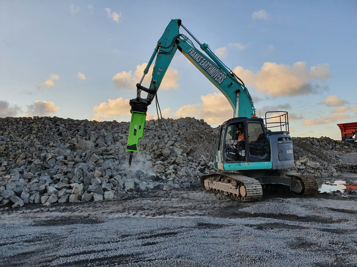 Excavator from Frank’s Earthmovers using an Impact Hydraulic Breaker attachment to break through large rocks at a construction site, set against a backdrop of a rocky landscape at sunset. The image emphasizes the power and effectiveness of the hydraulic breaker in heavy-duty rock-breaking tasks