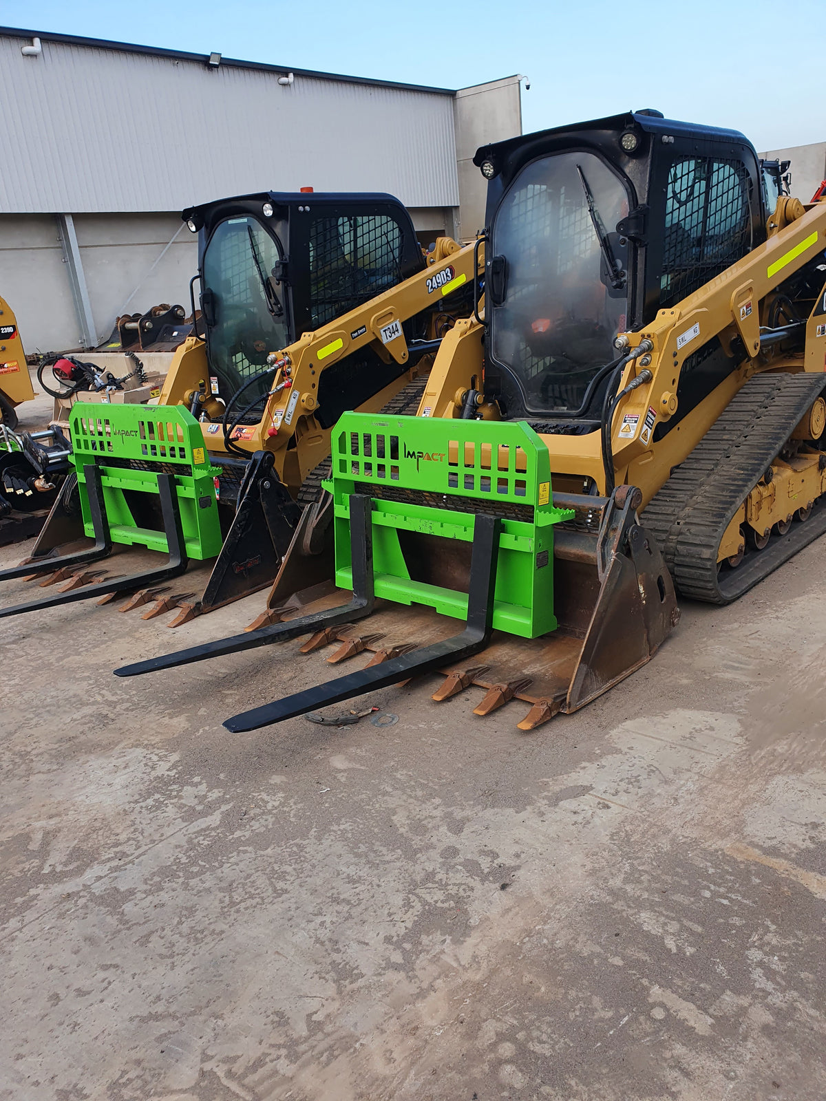 wo Caterpillar skid steer loaders equipped with green Impact pallet fork attachments, parked side by side in an outdoor industrial setting. The skid steers are prepared for heavy-duty lifting and material handling tasks.