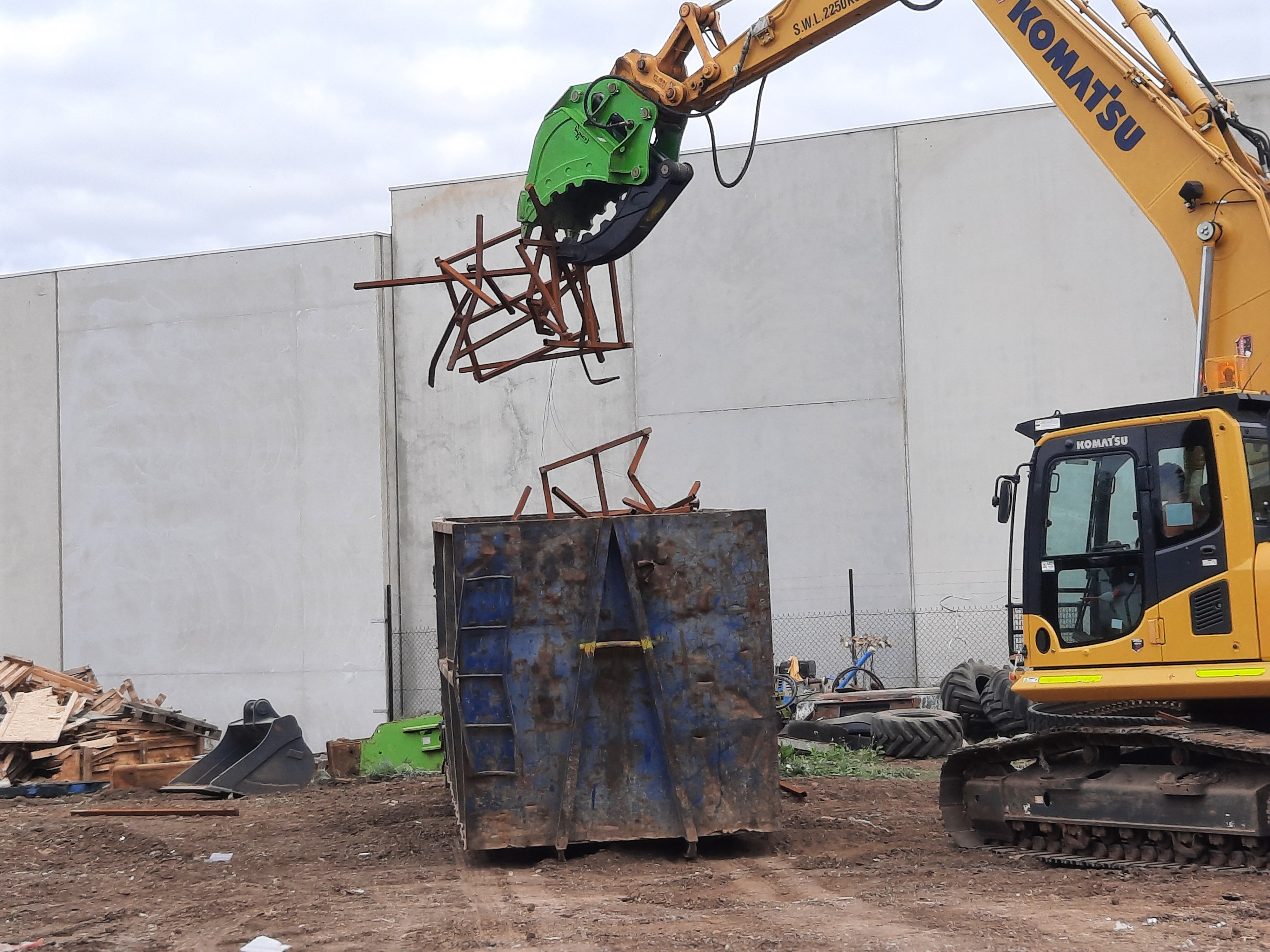 This image showcases a Komatsu excavator equipped with an Impact brand hydraulic grapple bucket attachment, efficiently cutting through metal structures on a demolition site. The hydraulic shear's powerful design allows for precise and efficient processing of steel and other materials, making it an essential tool for demolition, recycling, and scrap yard operations. This setup highlights the excavator's versatility and the shear's robust construction, optimized for heavy-duty tasks.