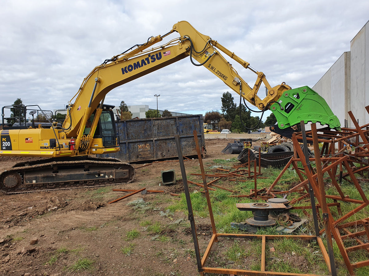 This image showcases a Komatsu excavator equipped with an Impact brand hydraulic grapple bucket attachment, efficiently cutting through metal structures on a demolition site. The hydraulic shear's powerful design allows for precise and efficient processing of steel and other materials, making it an essential tool for demolition, recycling, and scrap yard operations. This setup highlights the excavator's versatility and the shear's robust construction, optimized for heavy-duty tasks.