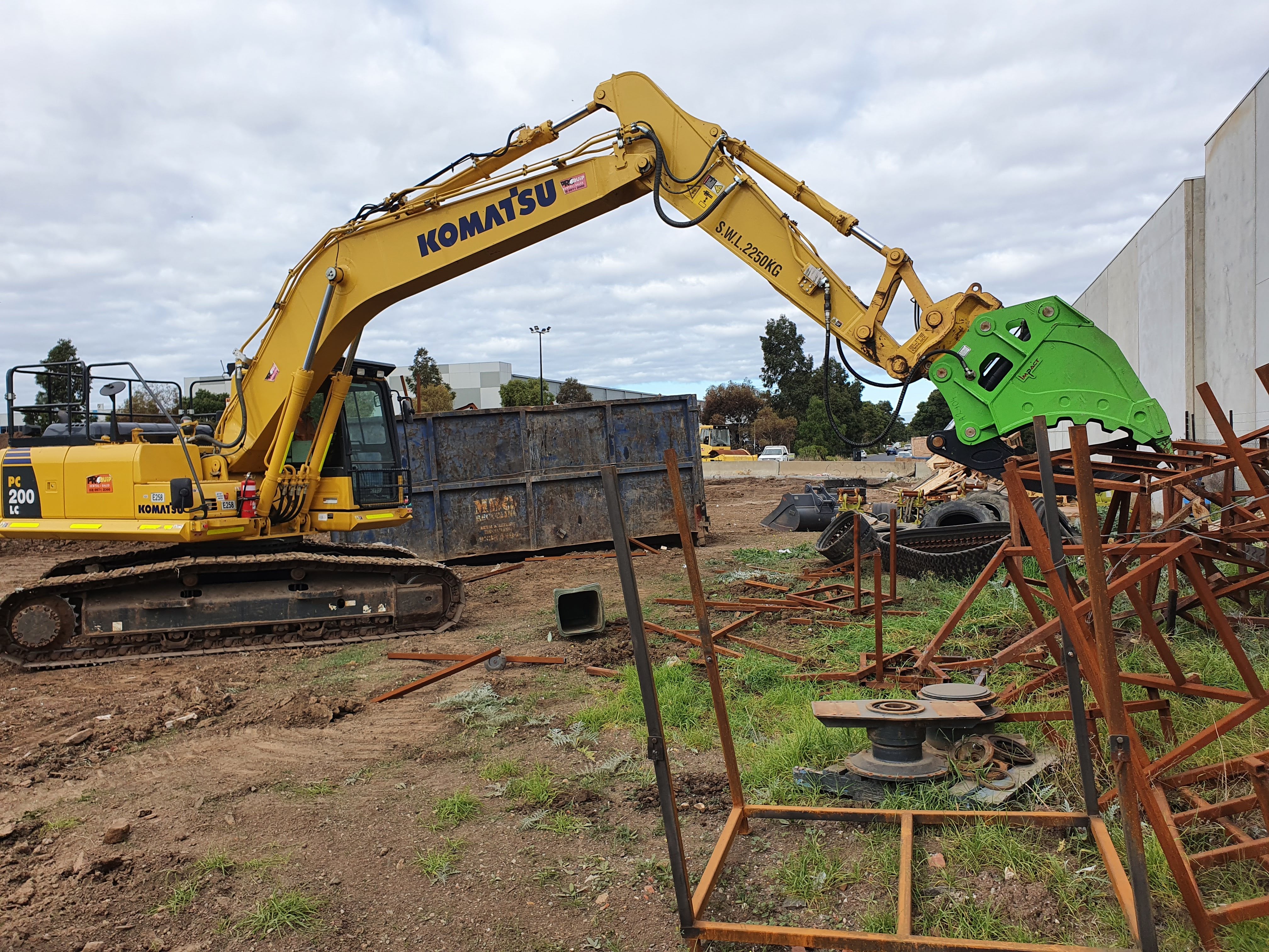 This image showcases a Komatsu excavator equipped with an Impact brand hydraulic grapple bucket attachment, efficiently cutting through metal structures on a demolition site. The hydraulic shear's powerful design allows for precise and efficient processing of steel and other materials, making it an essential tool for demolition, recycling, and scrap yard operations. This setup highlights the excavator's versatility and the shear's robust construction, optimized for heavy-duty tasks.