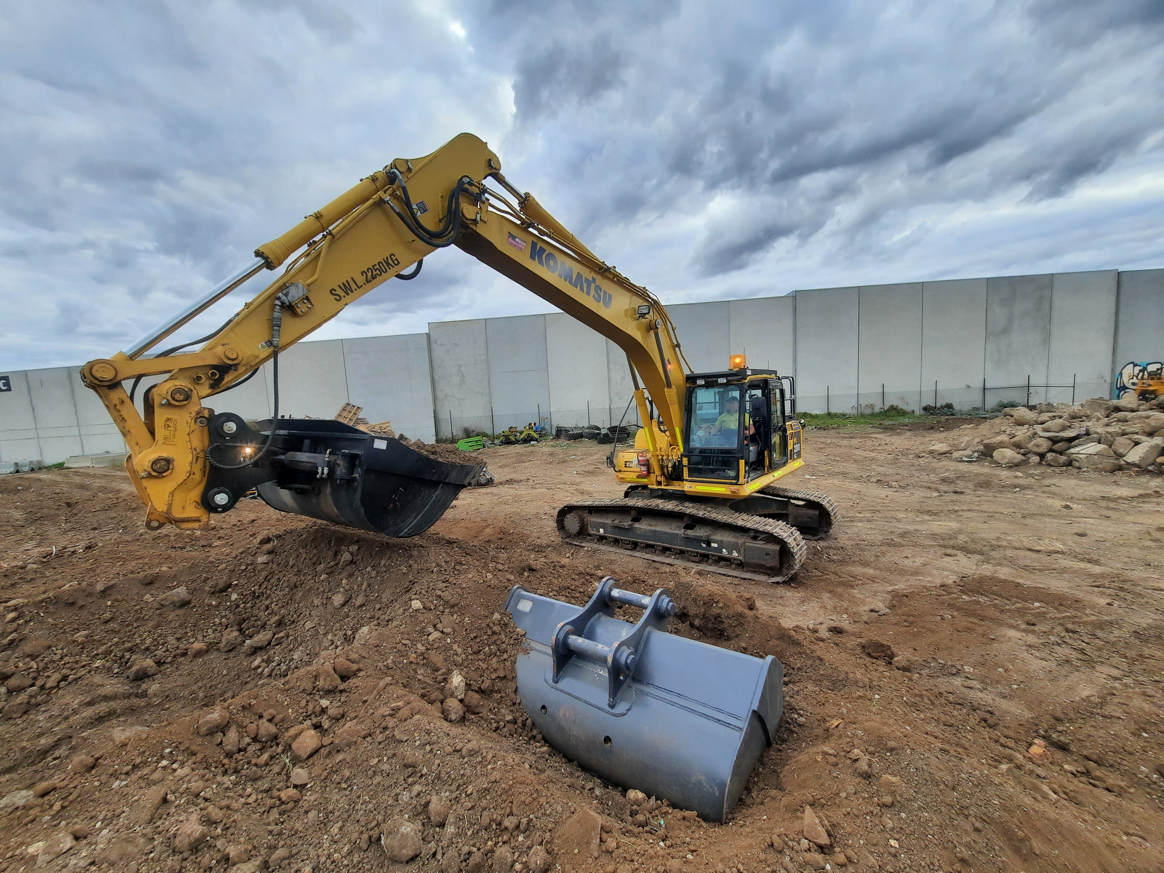 Komatsu excavator using an Impact bucket attachment to perform earthmoving tasks on a construction site. The bucket attachment showcases its versatility in digging and material handling, making it an essential tool for construction projects. A spare attachment sits nearby, highlighting the adaptability and efficiency of using Komatsu excavators with high-quality Impact attachments