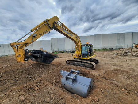 Komatsu excavator using an Impact bucket attachment to perform earthmoving tasks on a construction site. The bucket attachment showcases its versatility in digging and material handling, making it an essential tool for construction projects. A spare attachment sits nearby, highlighting the adaptability and efficiency of using Komatsu excavators with high-quality Impact attachments