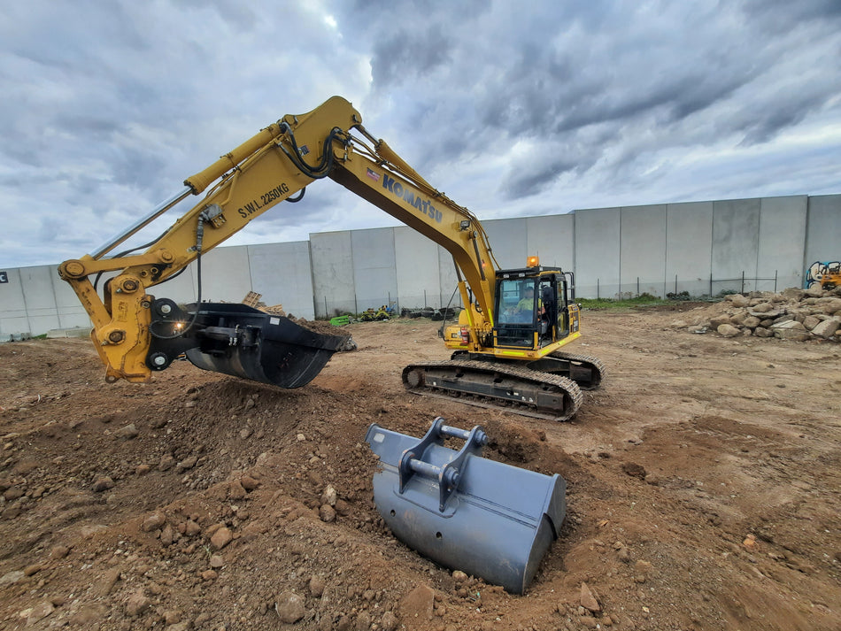 Komatsu excavator using an Impact bucket attachment to perform earthmoving tasks on a construction site. The bucket attachment showcases its versatility in digging and material handling, making it an essential tool for construction projects. A spare attachment sits nearby, highlighting the adaptability and efficiency of using Komatsu excavators with high-quality Impact attachments