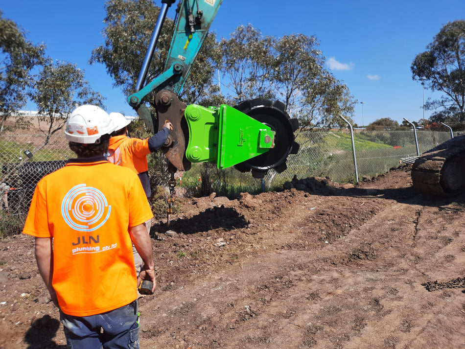 Workers on a construction site operating a Kobelco excavator equipped with an Impact Compaction Plate attachment, ideal for soil compaction and ground stabilization tasks. The compaction plate is highlighted in bright green, ensuring efficient and uniform compaction for infrastructure development.