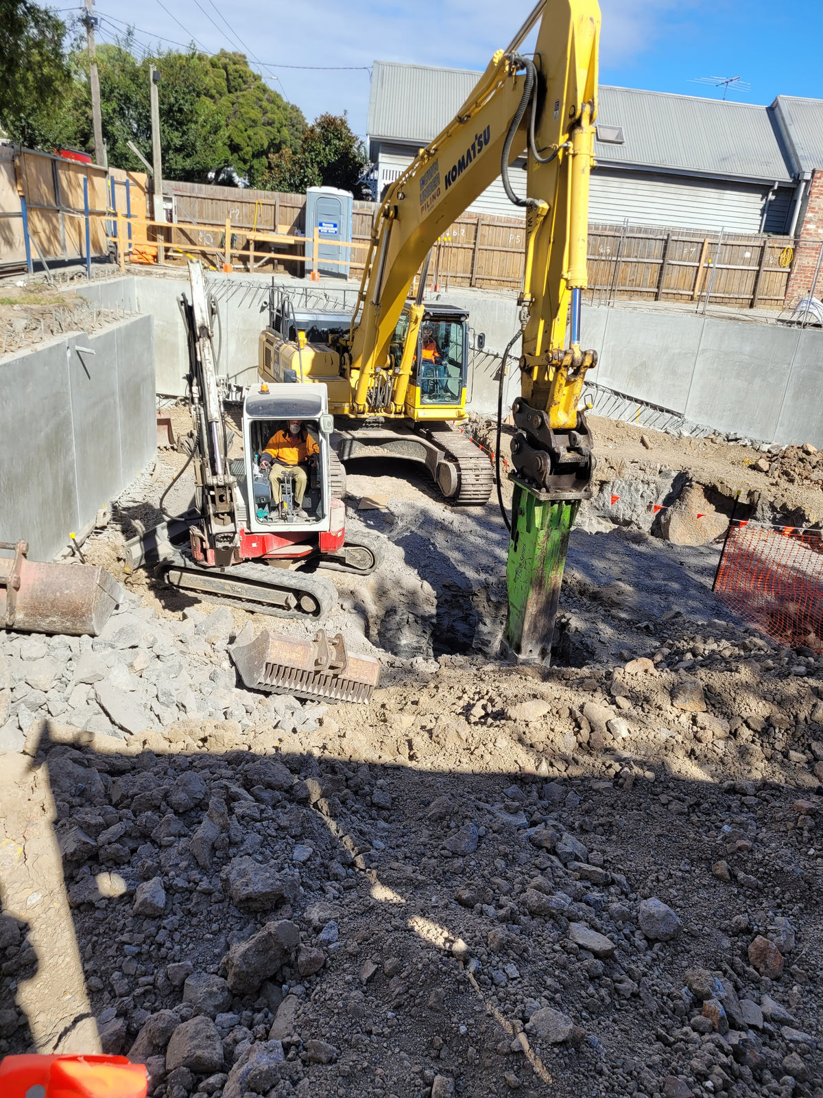 Excavation site featuring a Komatsu excavator equipped with an Impact hydraulic breaker and a mini excavator, performing rock-breaking tasks in a construction zone