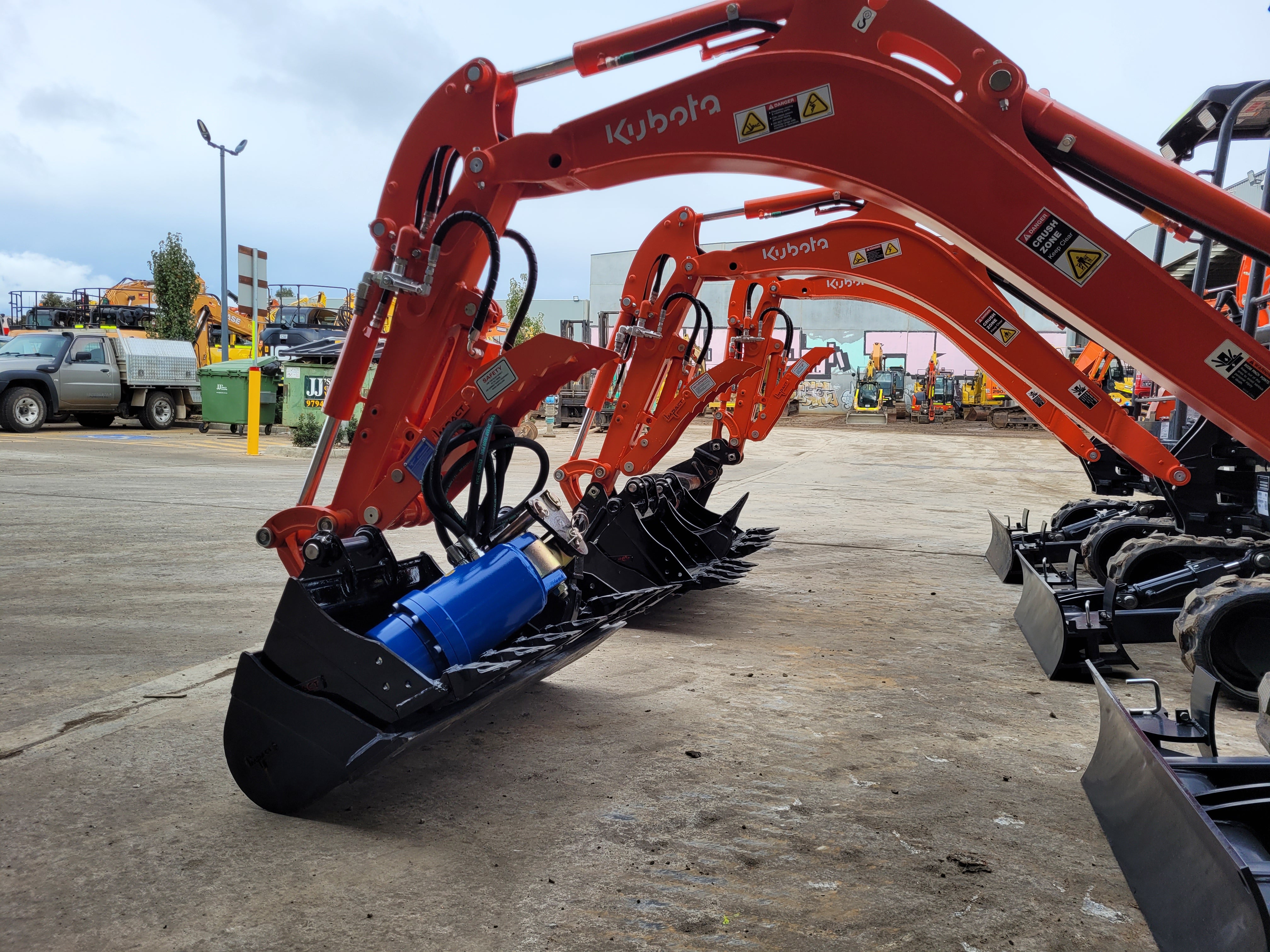 Row of Kubota mini excavators equipped with hydraulic thumbs and buckets, lined up in an industrial yard, ready for deployment in various construction, excavation, and landscaping projects. The hydraulic thumbs enhance material handling capabilities, making these machines ideal for precision work in tight spaces. Kubota mini excavators are known for their reliability, efficiency, and versatility on job sites