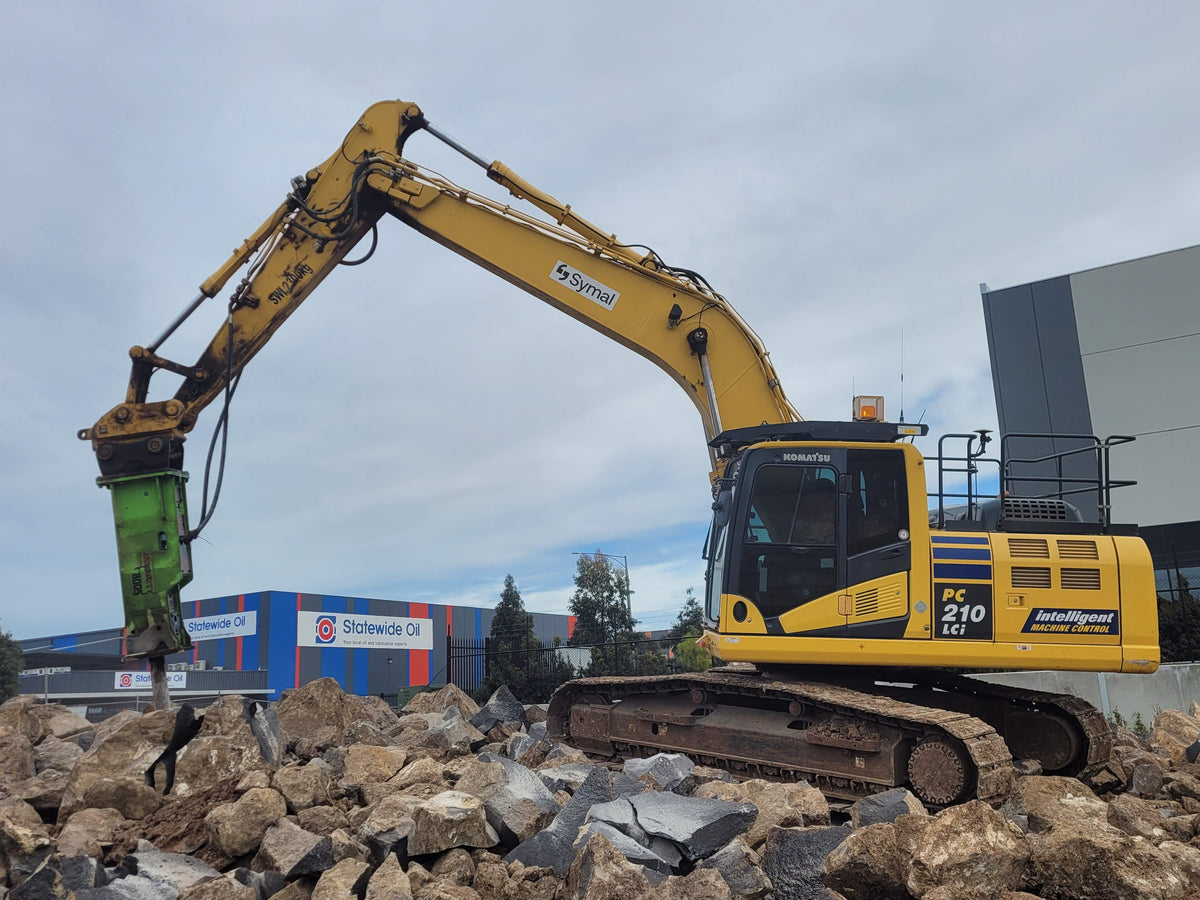 A Komatsu PC 210 LCi excavator equipped with a green Impact Hydraulic Breaker attachment, working on breaking large rocks at a construction site. The powerful hydraulic breaker is ideal for demolition and rock breaking tasks, ensuring efficient material removal. The excavator is positioned in front of an industrial facility, highlighting its heavy-duty capabilities
