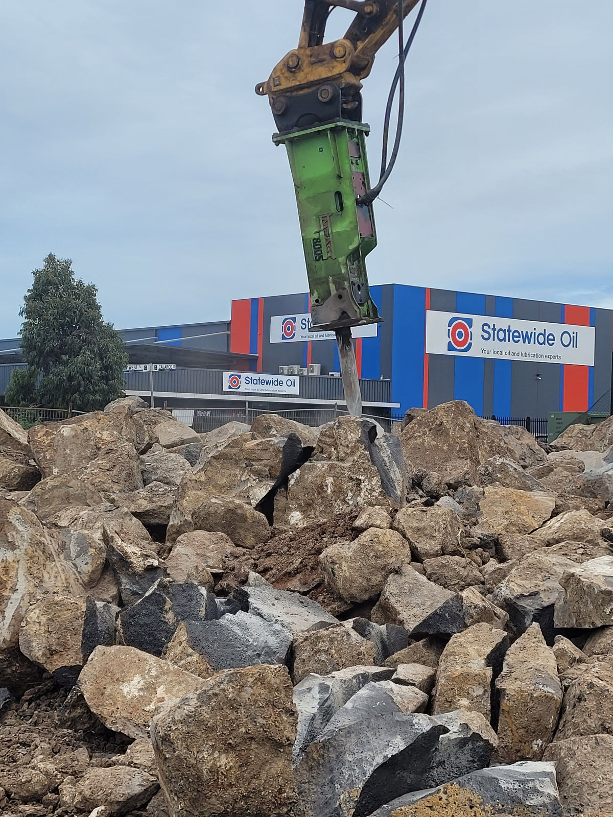 Impact Hydraulic Breaker attachment in action, mounted on an excavator and breaking through large rocks. The Statewide Oil building is visible in the background, highlighting the heavy-duty breaker’s application for industrial rock-breaking tasks at construction and demolition sites