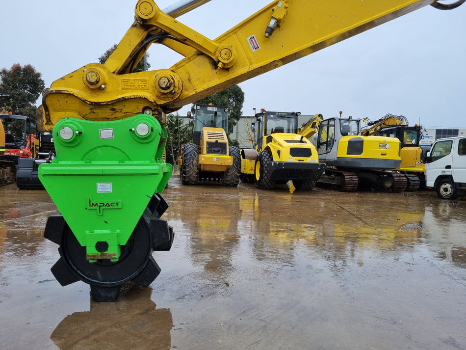 Close-up view of a bright green compaction wheel attachment connected to a yellow excavator arm, set against a wet construction yard. The reflection of the machinery is visible on the wet ground, and several other construction vehicles are parked in the background