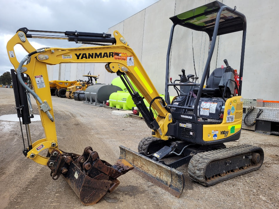 A yellow Yanmar mini excavator with a rusted attachment, parked on a gravel lot near other construction equipment, including a water tank and dump trucks. The excavator is equipped with a digging bucket and a dozer blade, highlighting its readiness for earthmoving tasks in an industrial setting