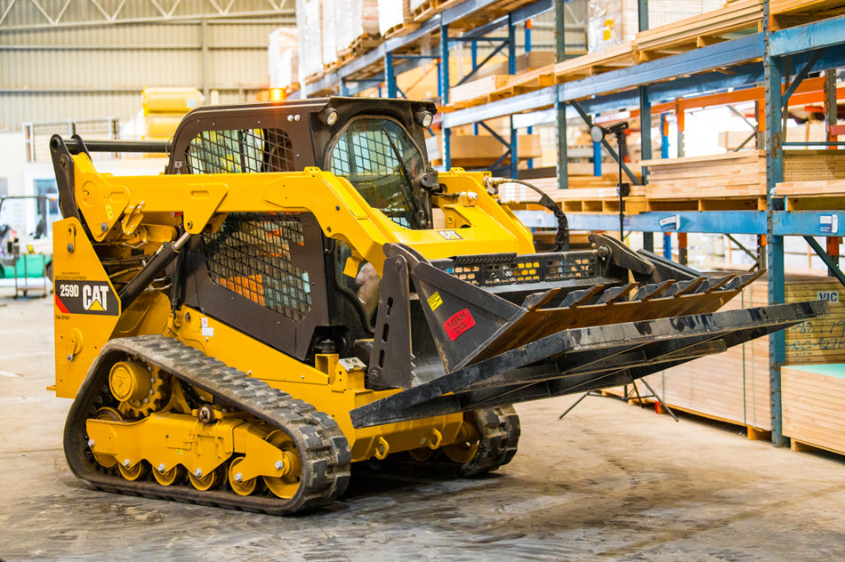 A yellow CAT 259D skid steer loader equipped with a 4-in-1 bucket and a smudge bar attachment inside a warehouse. The loader is positioned in front of shelving units filled with plywood sheets. The industrial environment is well-lit, showcasing the loader's robust design and the versatile attachments