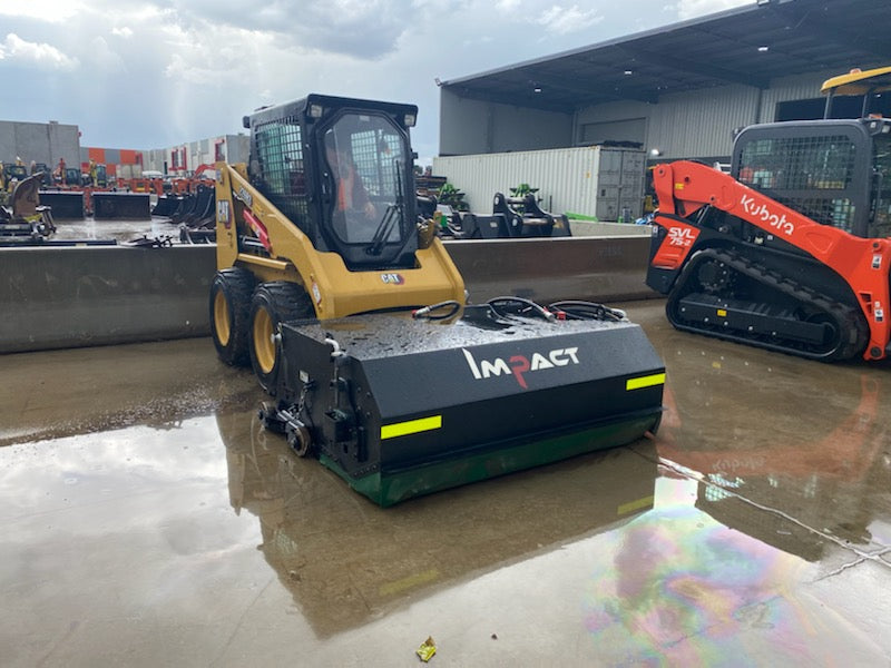 CAT 216B3 skid steer loader equipped with an Impact Attachments hydraulic broom, parked on a wet concrete surface outside a warehouse, ready for sweeping operations