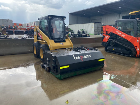 CAT 216B3 skid steer loader equipped with an Impact Attachments hydraulic broom, parked on a wet concrete surface outside a warehouse, ready for sweeping operations