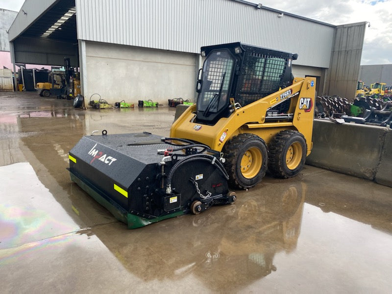 CAT 216B3 skid steer loader equipped with an Impact Attachments hydraulic broom, parked on a wet concrete surface outside a warehouse, ready for sweeping operations