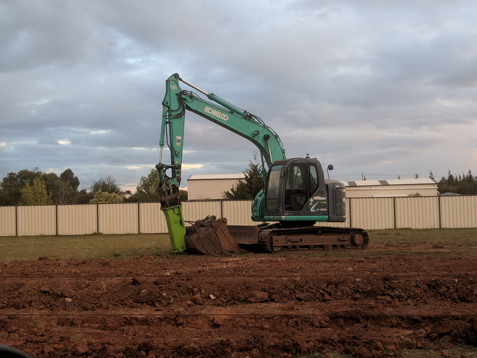 A Kobelco excavator working on a construction site, equipped with a green Impact Hydraulic Breaker attachment, designed for heavy-duty demolition tasks such as breaking up rock and concrete. The hydraulic breaker is a powerful tool for efficient excavation and material removal in construction projects
