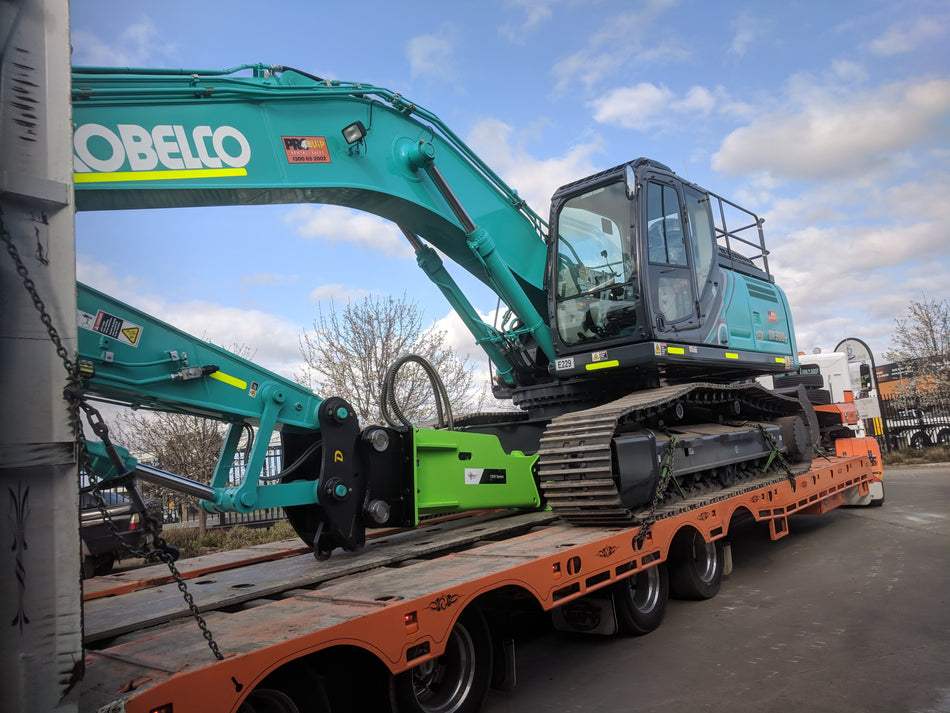 A Kobelco excavator loaded onto a flatbed truck, equipped with a bright green Impact Hydraulic Breaker attachment, ready for transport to a construction site. The hydraulic breaker is ideal for demolishing concrete, rock, and other hard materials, making it essential for construction and excavation projects