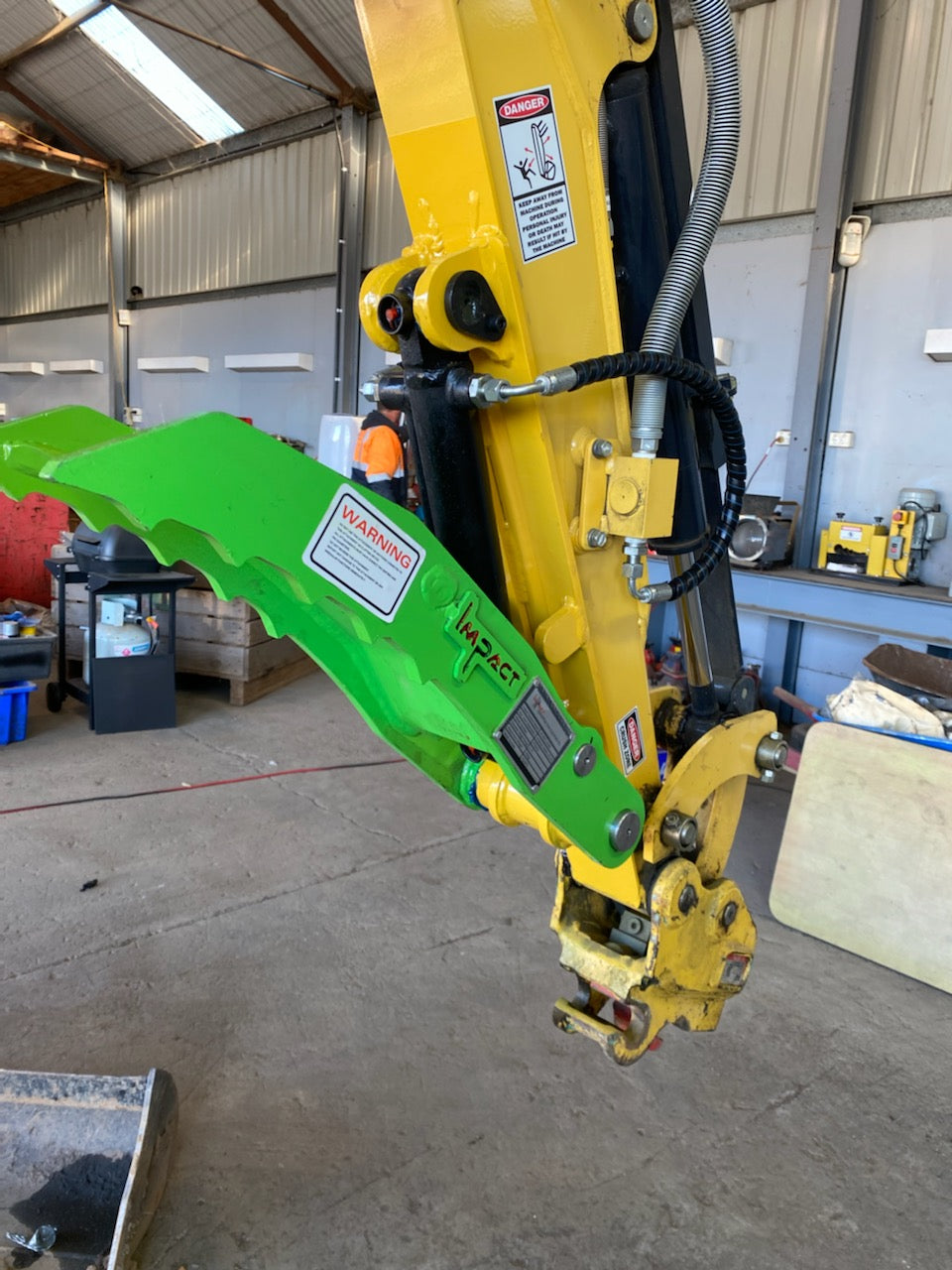 Close-up of a bright green Impact hydraulic thumb attached to a yellow excavator arm, inside an industrial workshop. The hydraulic thumb is designed for improved material handling, offering enhanced precision and grip for various excavation tasks. The setup showcases the durability and reliability of Impact attachments, making them a trusted choice for professionals in construction, demolition, and landscaping