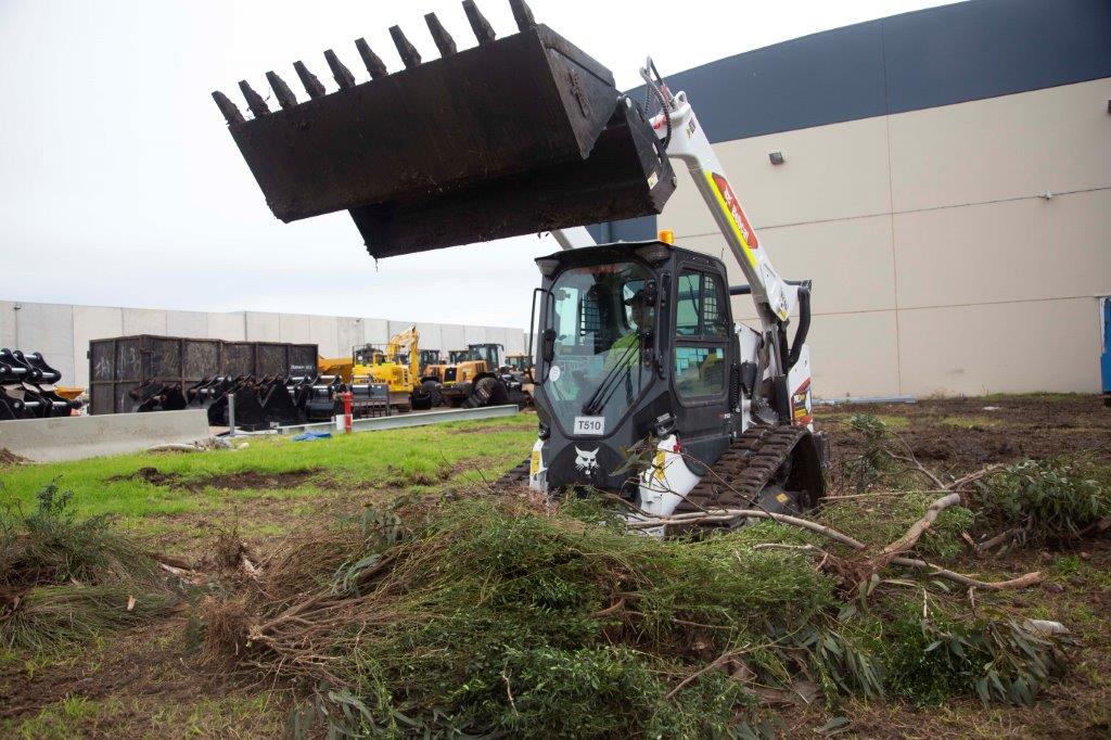 Track loader / skid steer equipped with a 4 in 1 bucket attachment, clearing brush and debris on a construction site, demonstrating versatility and power for heavy-duty tasks. Ideal for various excavation and earthmoving applications.