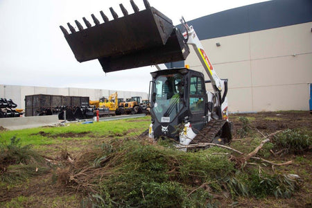 Track loader / skid steer equipped with a 4 in 1 bucket attachment, clearing brush and debris on a construction site, demonstrating versatility and power for heavy-duty tasks. Ideal for various excavation and earthmoving applications.