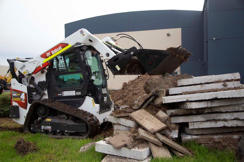 Track loader / skid steer equipped with a 4 in 1 bucket attachment, clearing brush and debris on a construction site, demonstrating versatility and power for heavy-duty tasks. Ideal for various excavation and earthmoving applications