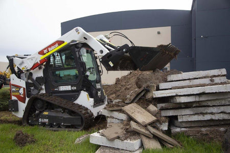 Track loader / skid steer equipped with a 4 in 1 bucket attachment, clearing brush and debris on a construction site, demonstrating versatility and power for heavy-duty tasks. Ideal for various excavation and earthmoving applications