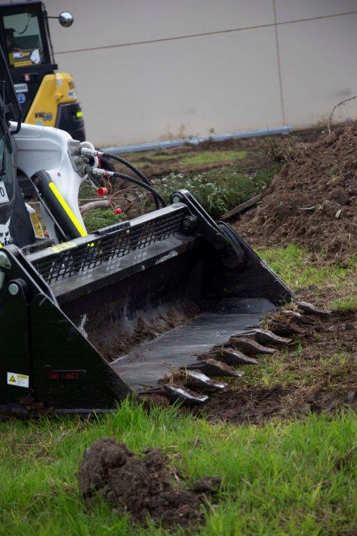 Track loader / skid steer equipped with a 4 in 1 bucket attachment, clearing brush and debris on a construction site, demonstrating versatility and power for heavy-duty tasks. Ideal for various excavation and earthmoving applications