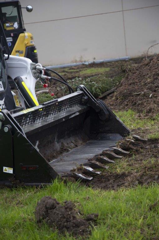 Track loader / skid steer equipped with a 4 in 1 bucket attachment, clearing brush and debris on a construction site, demonstrating versatility and power for heavy-duty tasks. Ideal for various excavation and earthmoving applications