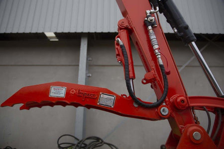 Close-up of an Impact Attachments hydraulic thumb in bright red, mounted on an excavator arm. The hydraulic thumb features a serrated edge designed for gripping and handling materials efficiently. The attachment is equipped with high-quality hydraulic fittings and hoses for optimal performance. Ideal for excavation and material handling tasks, this heavy-duty attachment showcases the durability and precision engineering of Impact Attachments.