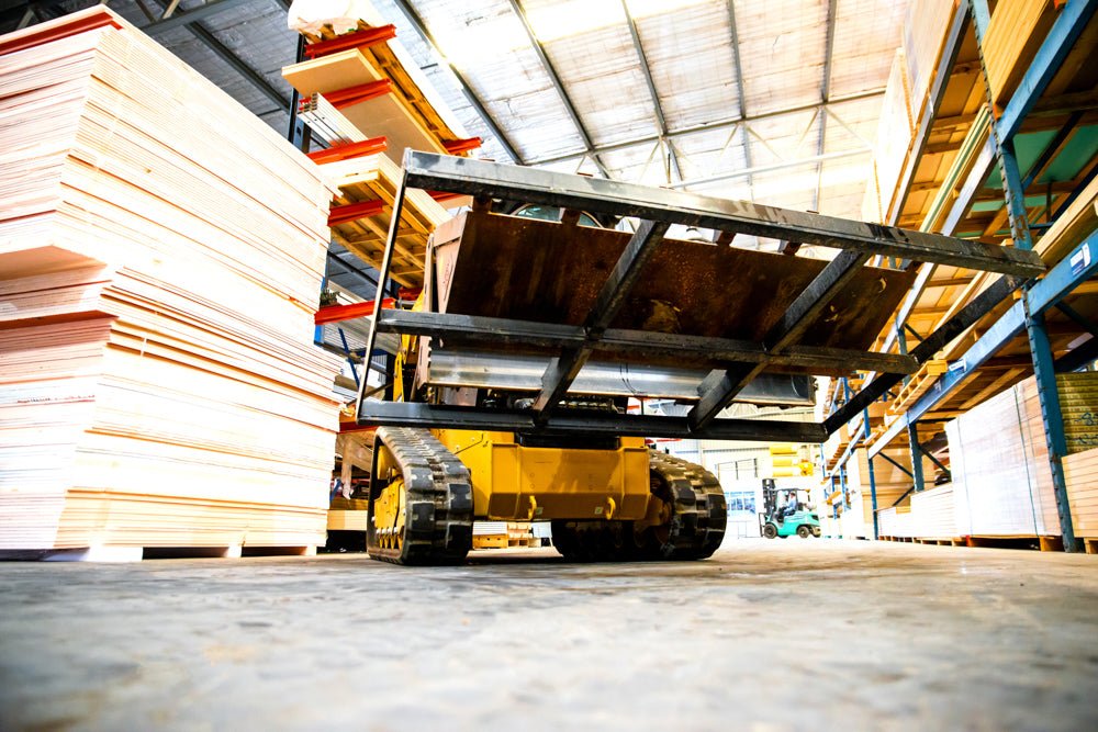 A low-angle view of a yellow skid steer loader equipped with a 4-in-1 bucket, lifting a large smudge bar inside a warehouse. The loader is moving through an aisle surrounded by stacks of plywood sheets and shelving. The perspective emphasizes the loader's treads and the underside of the raised smudge bar. The industrial setting is brightly lit with natural light filtering through the roof.