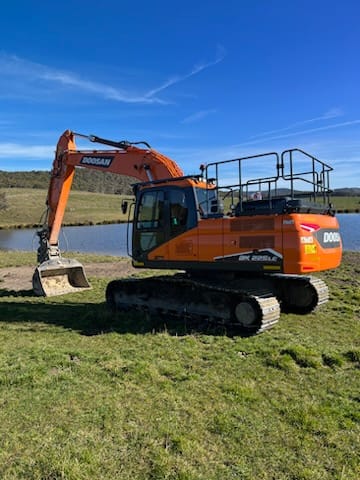 Doosan excavator on a grassy field near a water body under a clear blue sky, demonstrating the versatility and reliability of Doosan construction equipment in diverse terrains and conditions