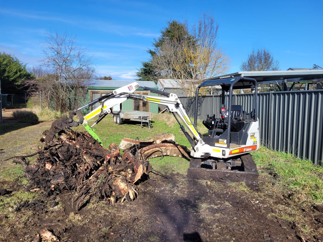 Bobcat mini excavator equipped with a hydraulic thumb attachment, clearing and moving large tree stumps and roots in a backyard setting, ideal for landscaping and heavy-duty yard work
