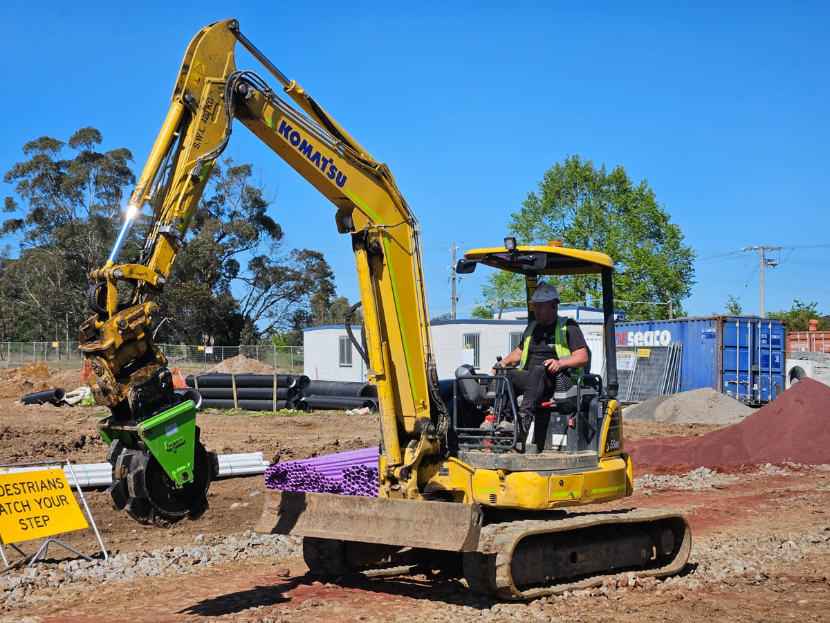 Komatsu excavator operated by a worker on a construction site, equipped with a green hydraulic attachment, with a sign reading 'PEDESTRIANS WATCH YOUR STEP' in the foreground, and construction materials in the background.