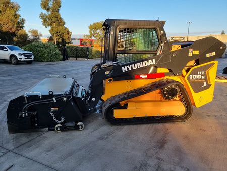 Hyundai HT100V skid steer loader with an Impact Attachments hydraulic broom in operation on a concrete surface, preparing for sweeping tasks at an industrial site during early morning light.