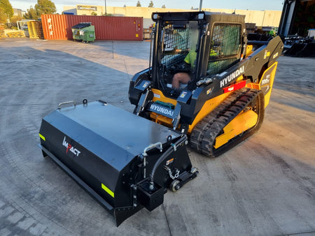 Hyundai HT100V skid steer loader with an Impact Attachments hydraulic broom in operation on a concrete surface, preparing for sweeping tasks at an industrial site during early morning light.
